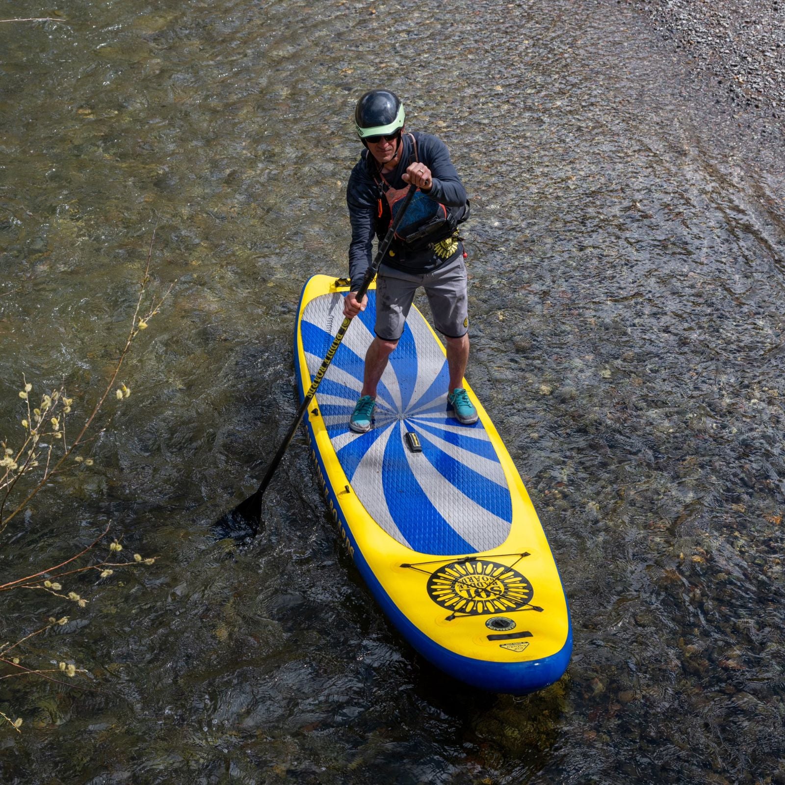 A person paddling on a yellow and blue SOLrey inflatable paddle board in a shallow, clear river, wearing a helmet, sunglasses, and a life jacket.