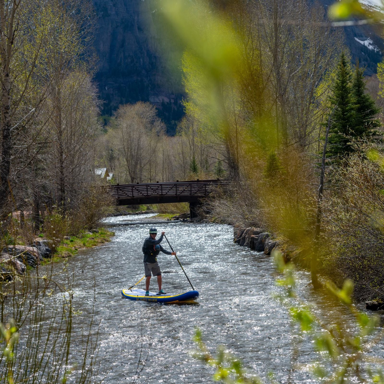 A paddler on a SOLrey inflatable paddle board gliding through a scenic river with a bridge and mountainous background, surrounded by trees.