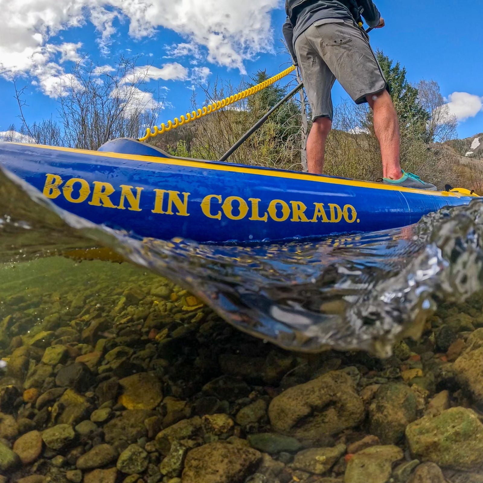 Close-up shot of the side of the SOLrey inflatable paddle board in the water, showing the text "Born in Colorado" with a person standing on it, paddling.